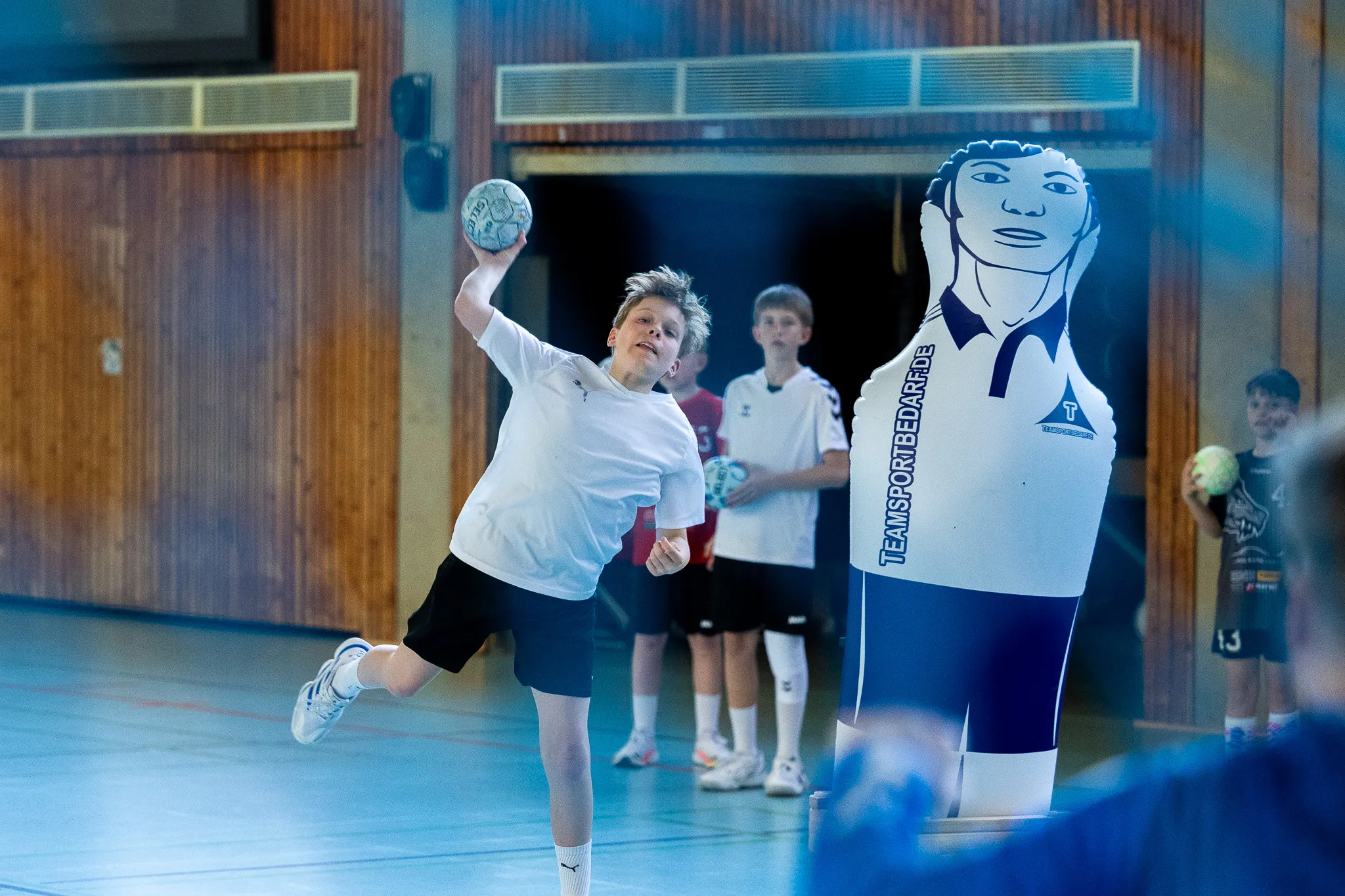 Ein Jugendlicher wirft im Hallensport einen Handball, während andere Kinder im Hintergrund stehen. Rechts steht eine aufblasbare Figur mit dem Schriftzug „teamsportbedarf.de“.