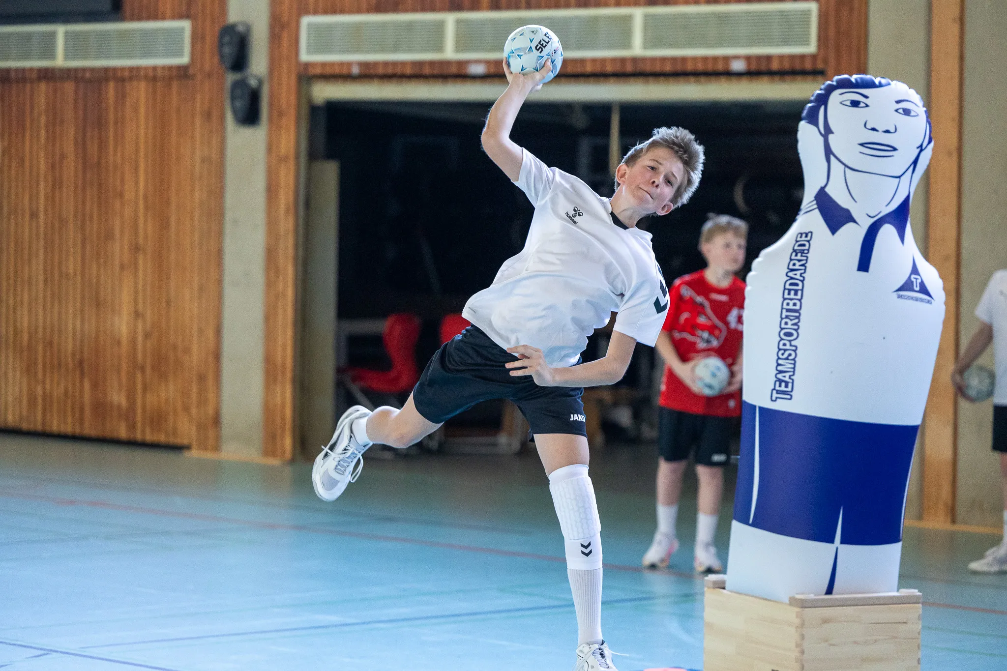 Ein Jugendlicher wirft beim Handball in einer Sporthalle und springt dabei in die Luft. Im Hintergrund stehen weitere Kinder und ein aufblasbarer Torwart.