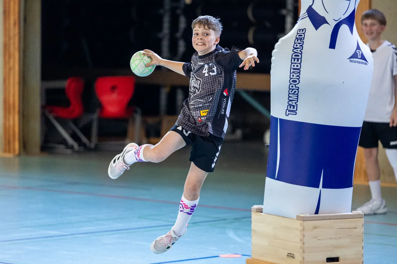 Ein Jugendlicher wirft im Handballspiel einen Ball, während er in der Luft springt. Im Hintergrund steht eine aufblasbare Figur mit der Aufschrift „Teamsportbedarf.de“.