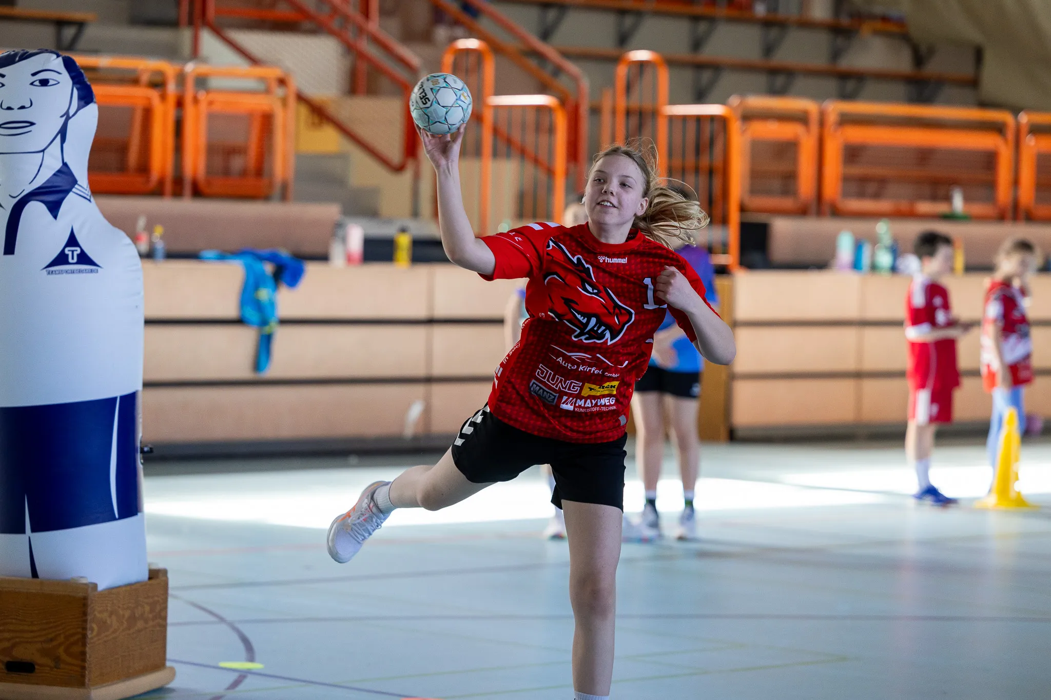 Eine junge Handballspielerin in rotem Trikot wirft den Ball während eines Trainings in einer Sporthalle. Im Hintergrund weitere Kinder beim Spiel.