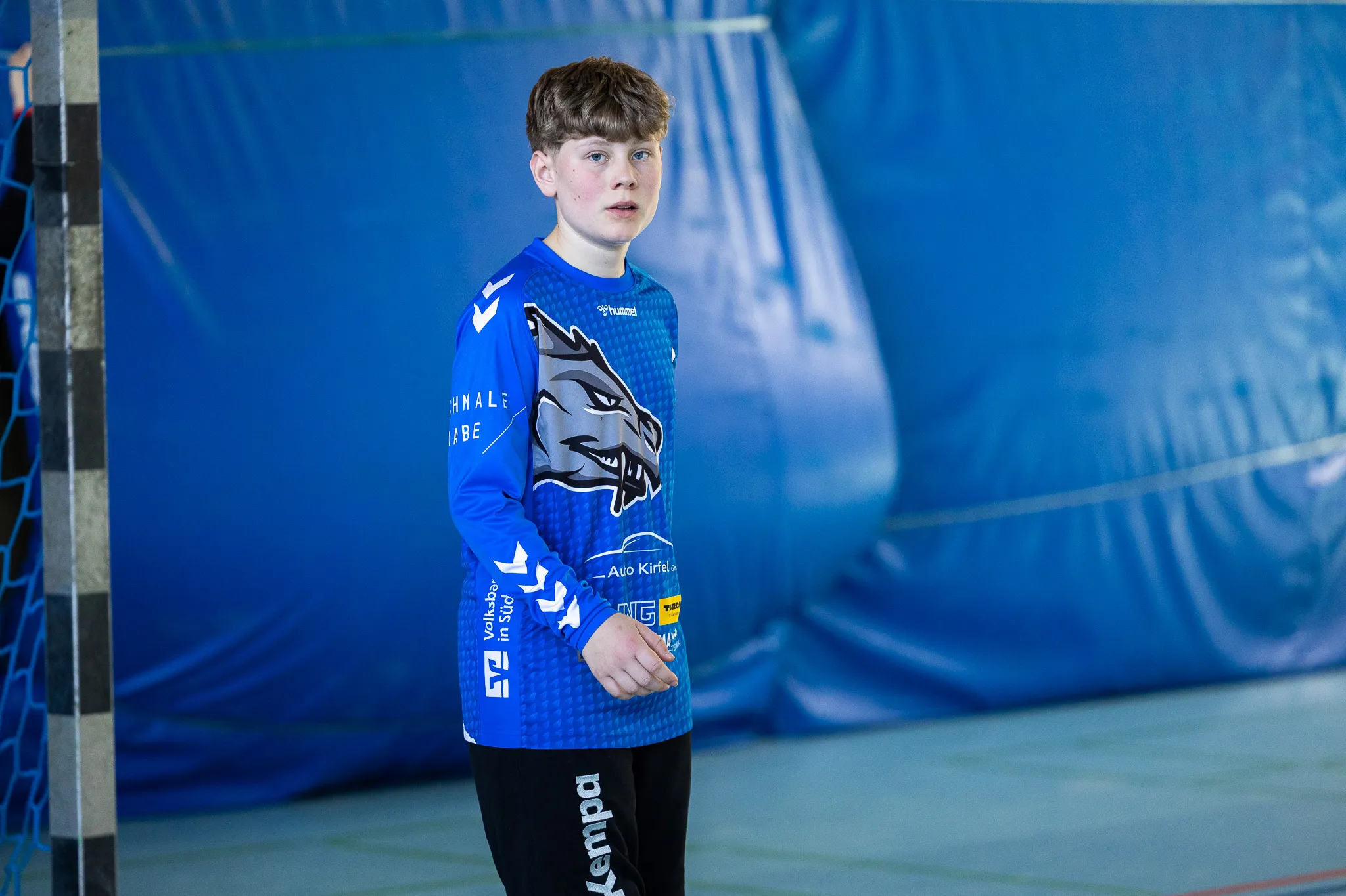 Ein Jugendlicher in Handballbekleidung steht auf einem roten Spielfeld vor blauem Hintergrund und blickt konzentriert nach links.