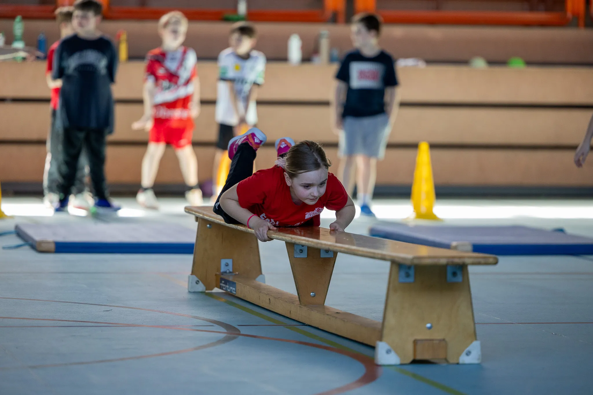 Ein Mädchen in roter Sportkleidung balanciert auf einem schmalen Holzbalken in einer Turnhalle, während andere Kinder zusehen.