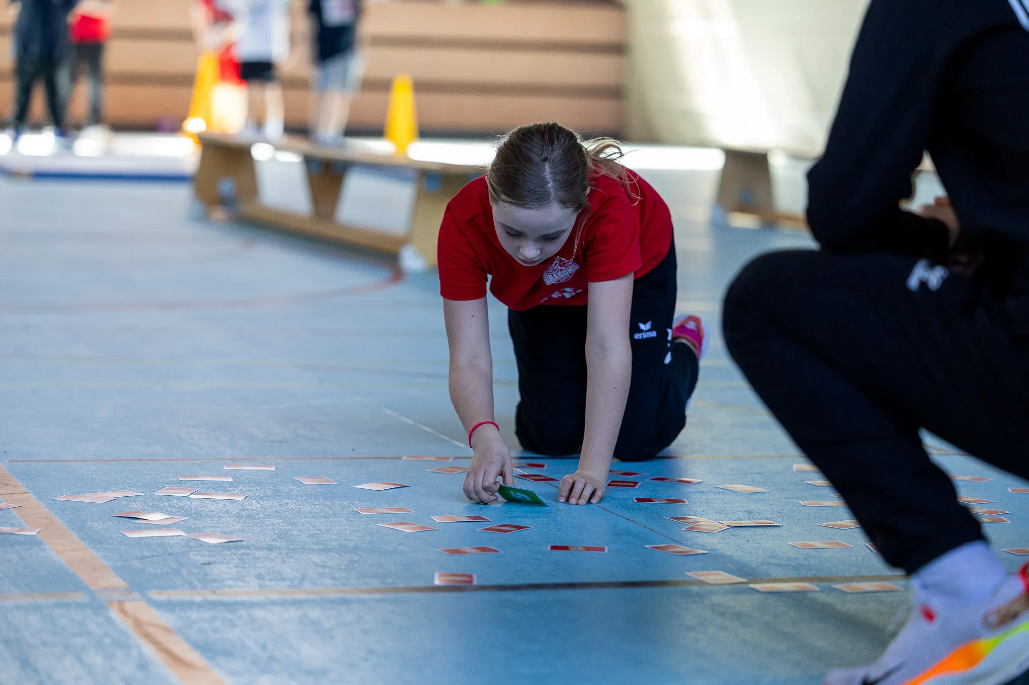 Ein Mädchen in rotem Trainingsanzug hockt auf dem Boden einer Sporthalle und legt Karten aus. Im Hintergrund sind weitere Kinder und Sportgeräte zu sehen.
