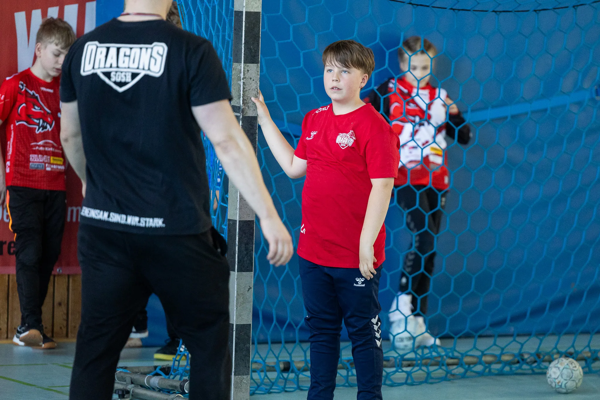 Ein Junge im roten Trikot steht vor einem Handballtor und deutet mit der Hand nach oben. Im Hintergrund trainieren weitere Kinder mit einem Betreuer.
