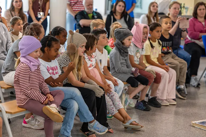 Eine Gruppe von Kindern in Kostümen und mit Gesichtsbemalung sitzt auf Bänken während einer Theateraufführung.