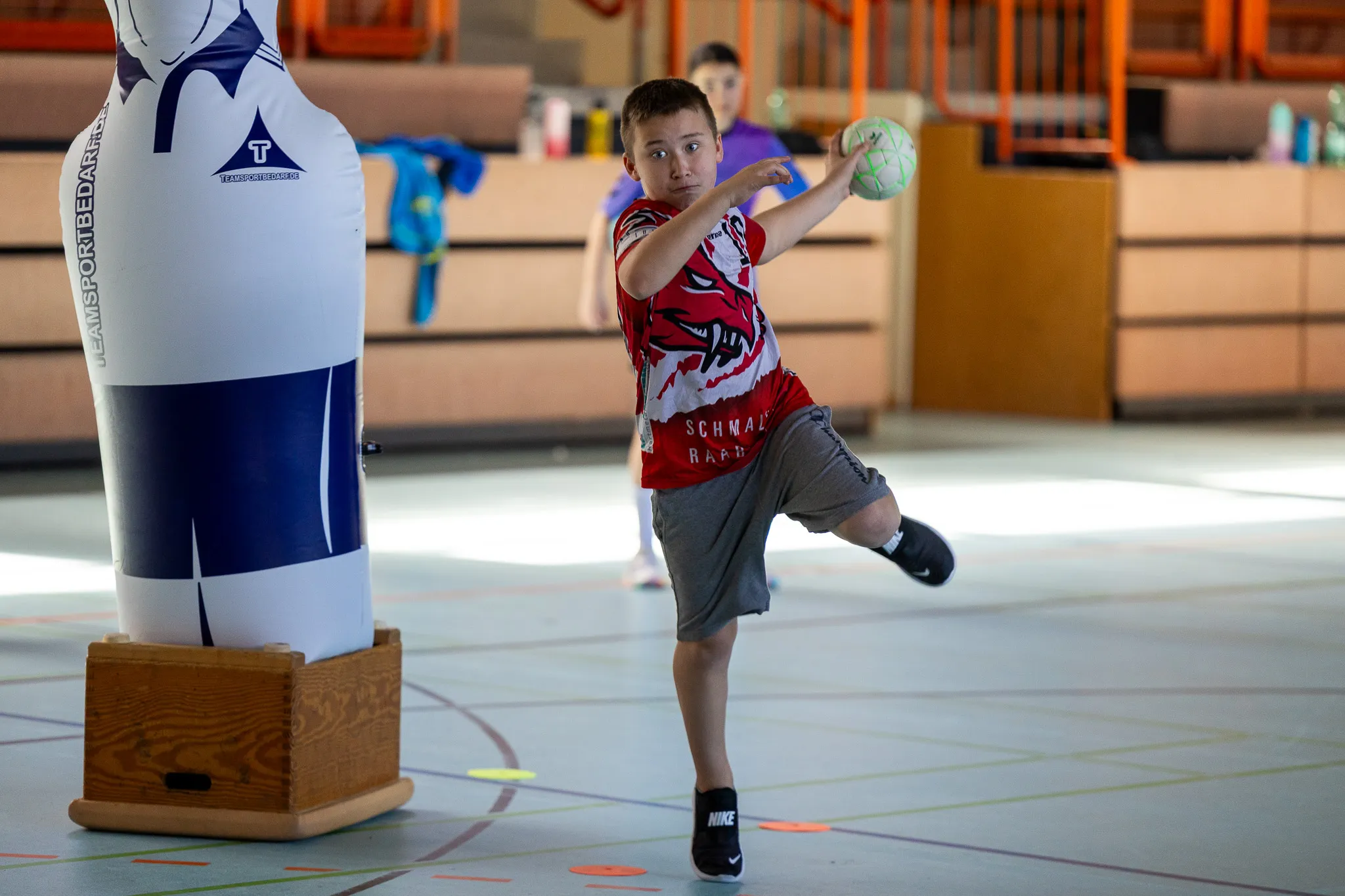 Ein Junge wirft einen Handball in einer Sporthalle. Im Hintergrund steht ein aufblasbarer Torwart und ein weiteres Kind.