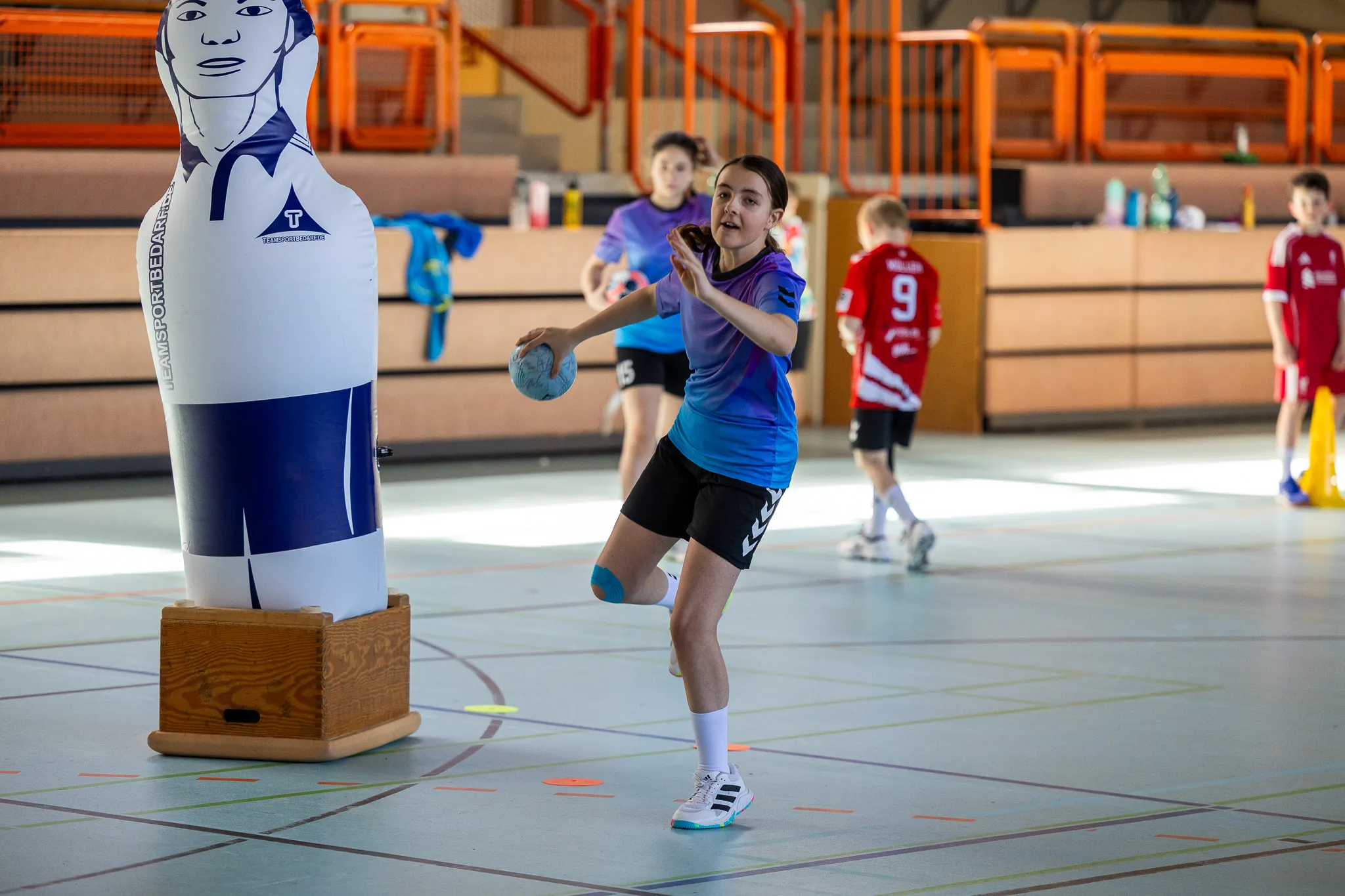 Eine junge Handballspielerin wirft den Ball während eines Trainings in einer Sporthalle. Im Hintergrund steht eine aufblasbare Figur eines Handballspielers und weitere Kinder.