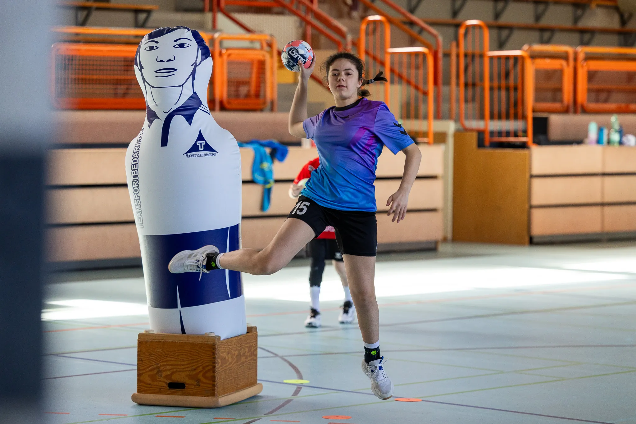 Eine junge Handballspielerin springt in einer Sporthalle in die Luft, um den Ball zu werfen. Neben ihr steht eine aufblasbare Figur eines Handballspielers.