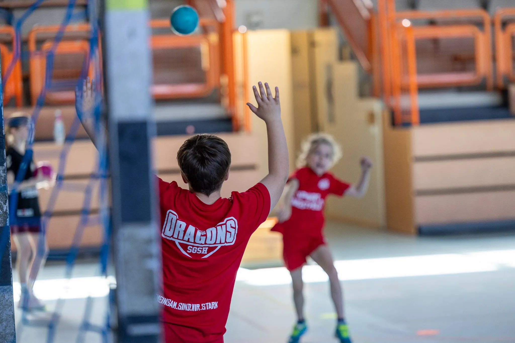 Ein Kind mit dem Trikot der „Dragons SGSH“ versucht, einen Ball in der Sporthalle zu werfen, während ein anderes Kind im Hintergrund darauf wartet.