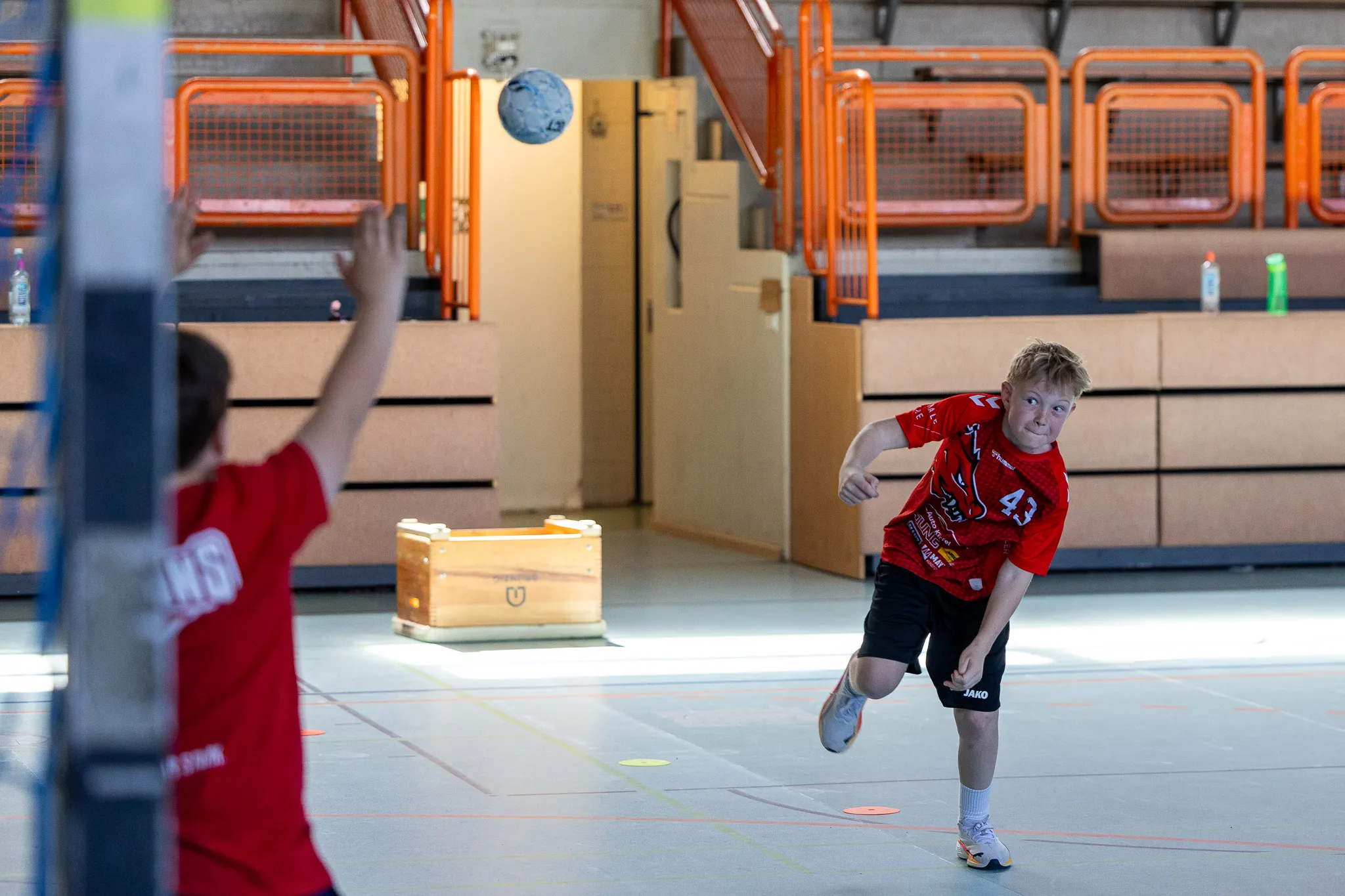 Ein junger Handballspieler in rotem Trikot mit der Nummer 43 wirft den Ball während eines Trainings in einer Sporthalle. Im Hintergrund ein weiterer Spieler mit erhobener Hand.