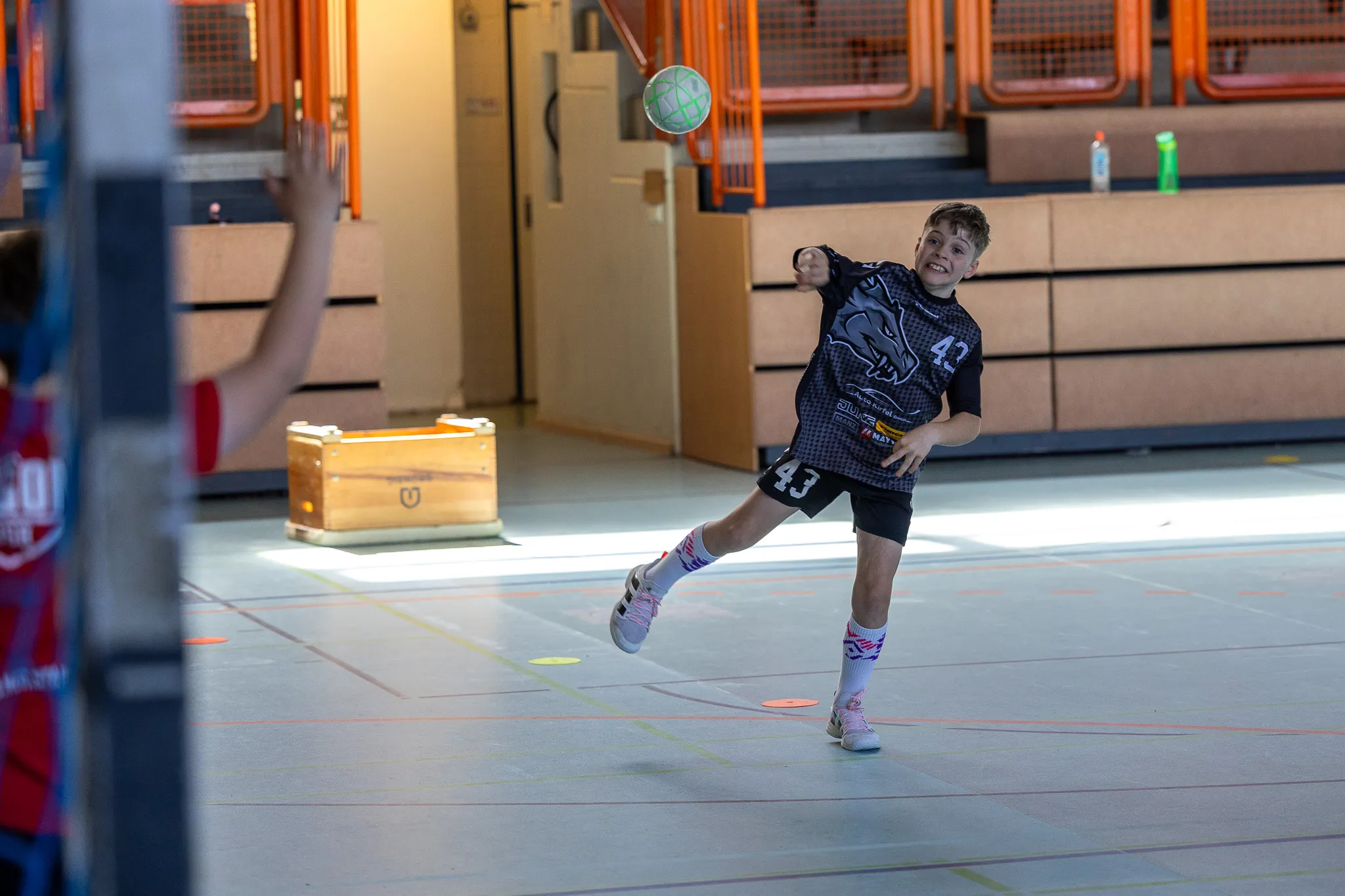 Ein Jugendlicher in Handballbekleidung wirft den Ball während eines Trainings in einer Sporthalle. Im Hintergrund ein weiterer Spieler und orangefarbene Zuschauertribünen.