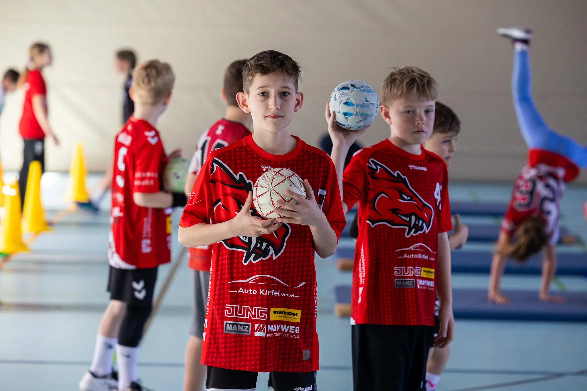Ein Jugendlicher im Handballtrikot hält einen Handball vor sich, während im Hintergrund andere Kinder trainieren und eine Athletin Dehnübungen macht.
