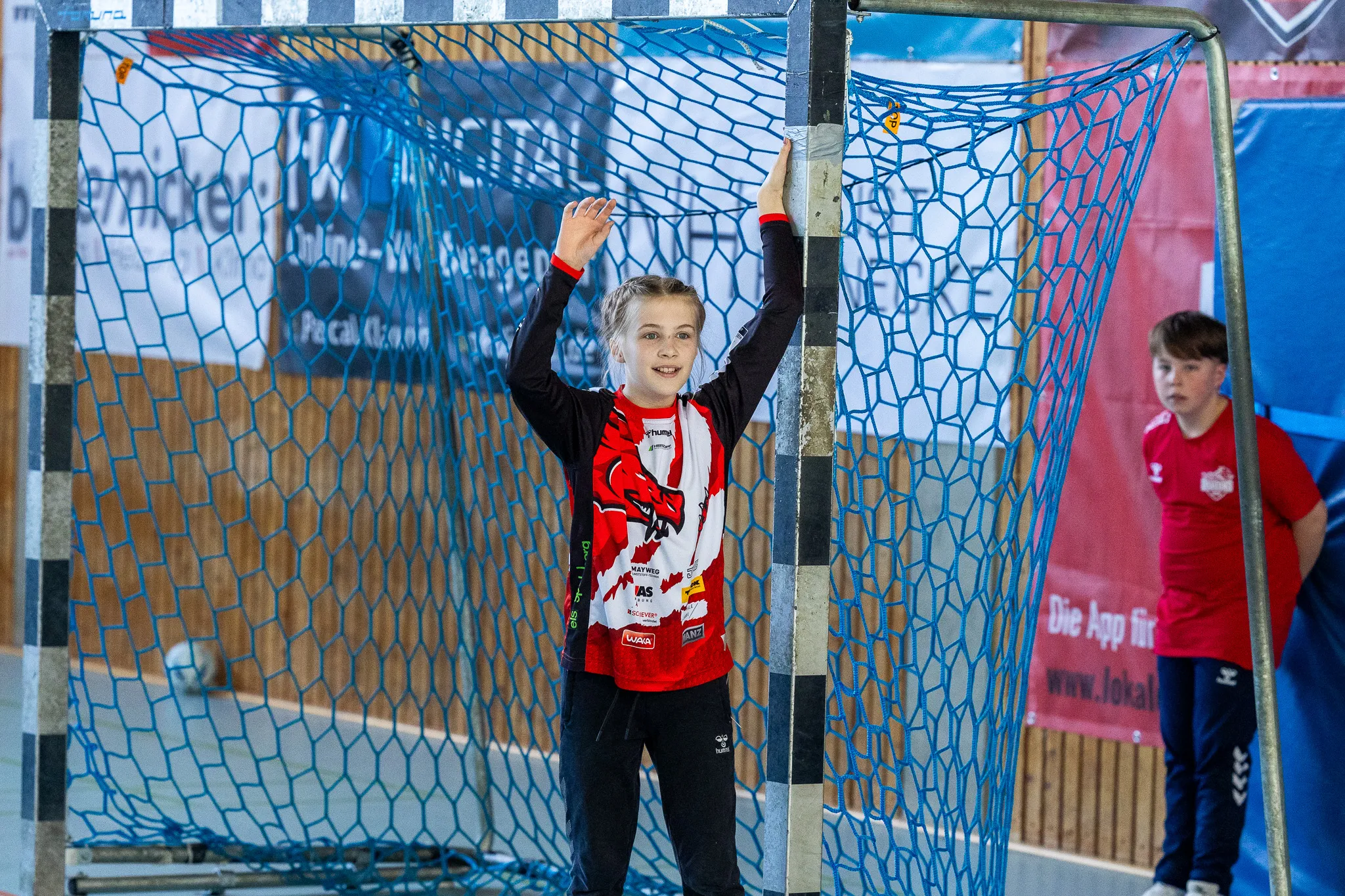 Junge Handballspielerin steht vor dem Tor und streckt eine Hand nach oben, während ein Teamkollege im Hintergrund steht.