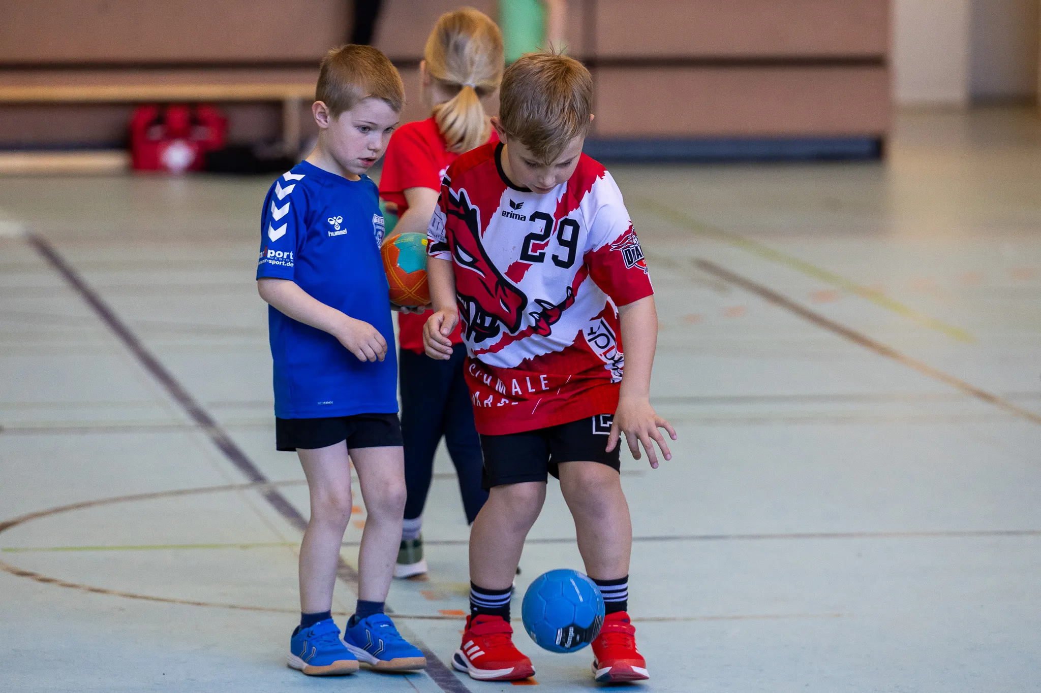 Zwei junge Handballspieler trainieren in einer Sporthalle. Ein Spieler in rotem Trikot mit der Nummer 29 dribbelt den Ball, während ein anderer ihn in blauem Trikot unterstützt.