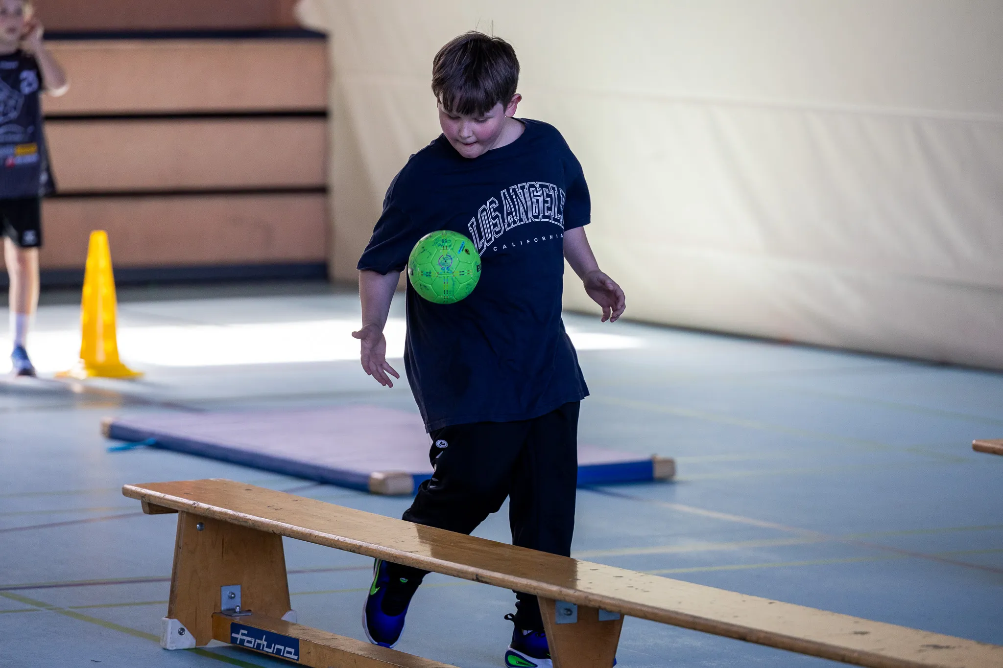 Ein Junge balanciert mit einem Ball auf einem schmalen Balken in einer Turnhalle, während andere Kinder im Hintergrund trainieren.