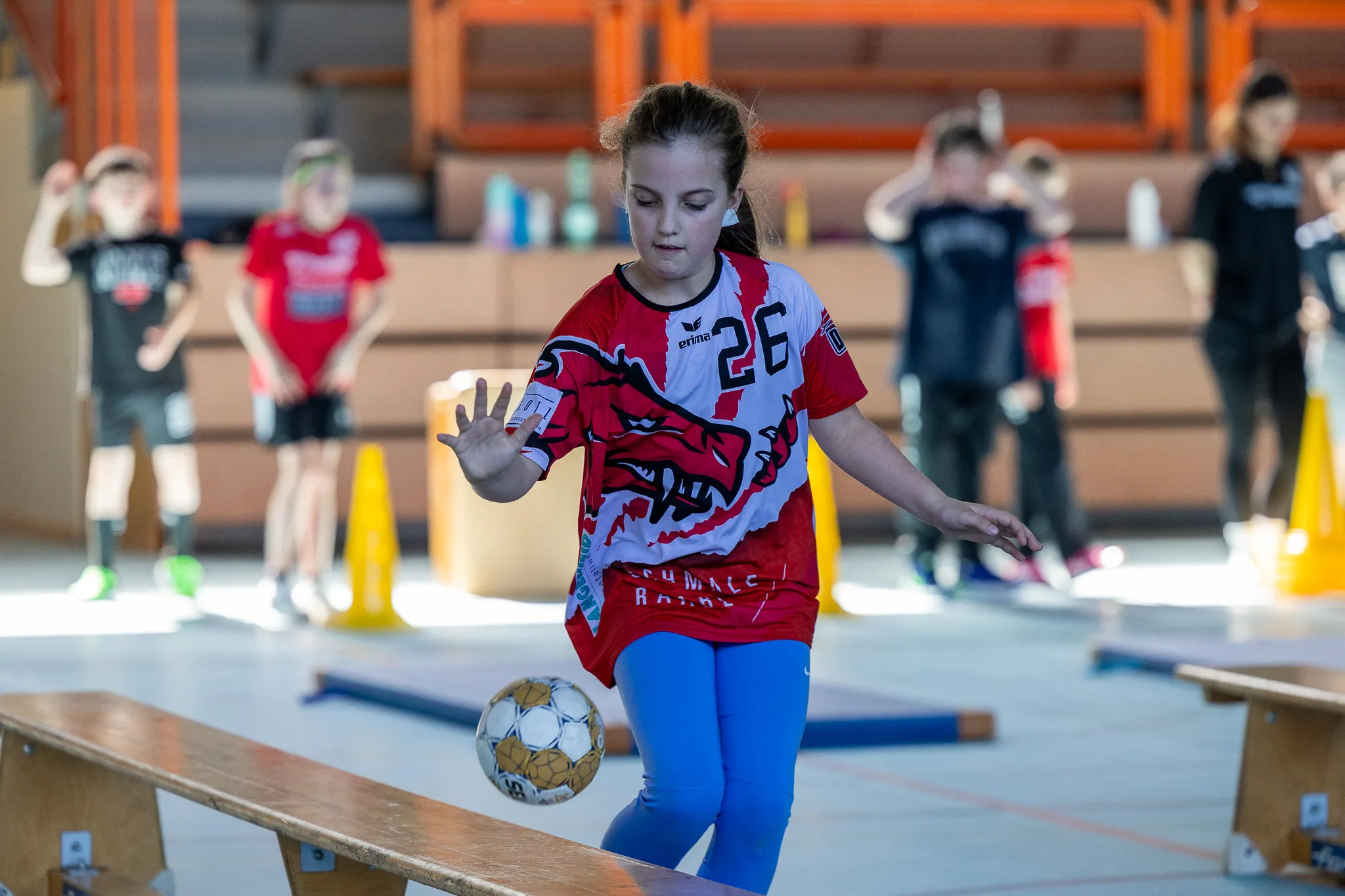 Ein Mädchen in Handballkleidung dribbelt einen Ball in einer Sporthalle. Im Hintergrund stehen weitere Kinder und trainieren.