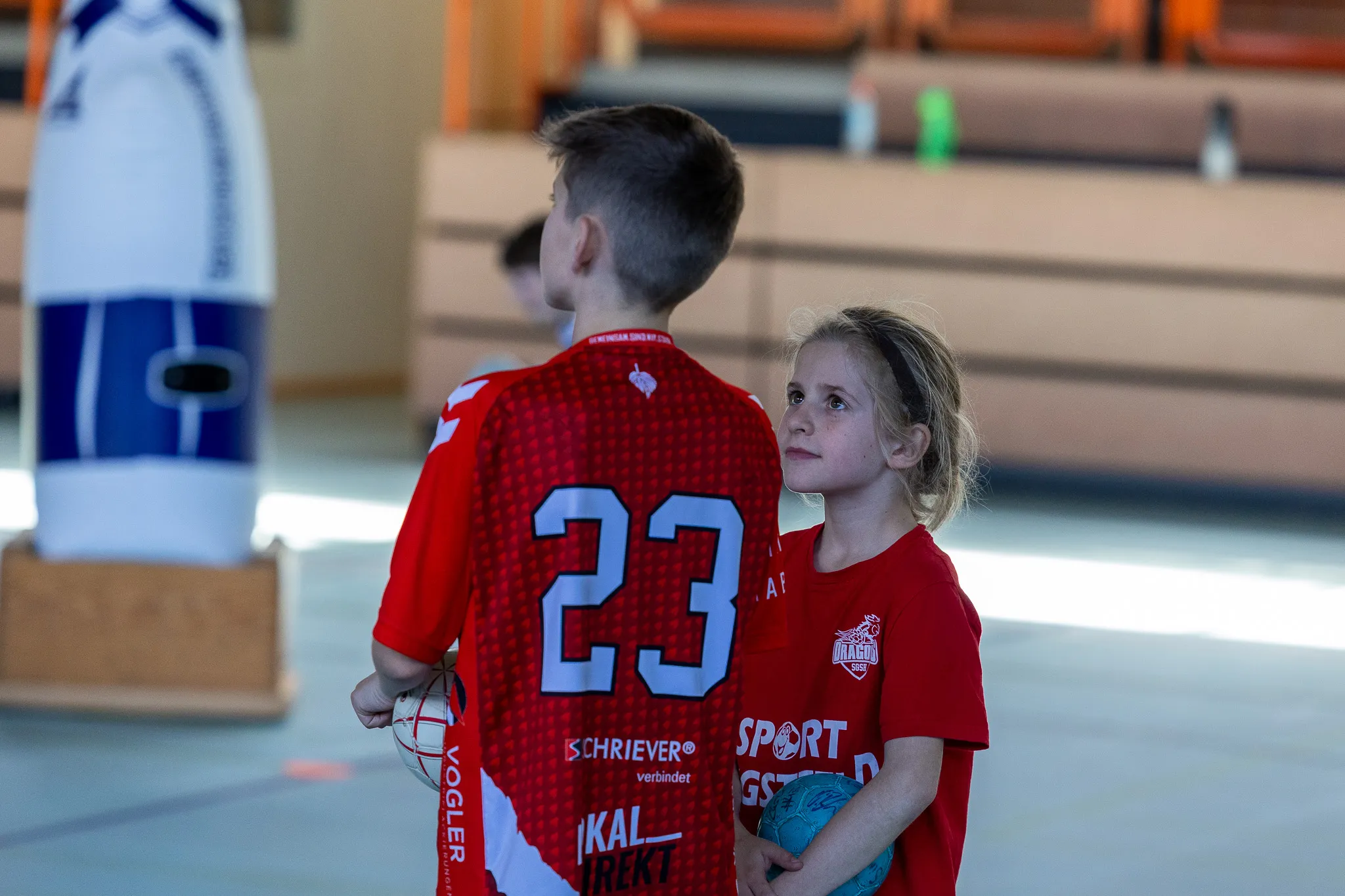 Junge Handballspielerin mit rotem Trikot und der Nummer 23 hält einen Ball und blickt konzentriert nach vorne, während ein Teamkollege im Hintergrund steht.