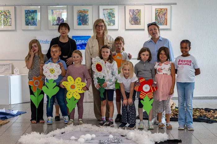 Zehn Kinder und drei Erwachsene posieren in einem Raum, wobei einige Kinder große, bunte Papierblumen halten.