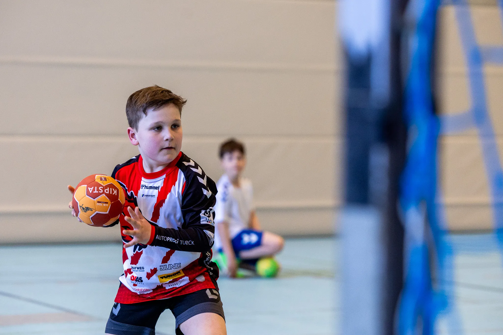 Ein junger Handballspieler in rotem Trikot mit dem Ball, konzentriert blickend zur Seite während eines Trainings in einer Sporthalle.