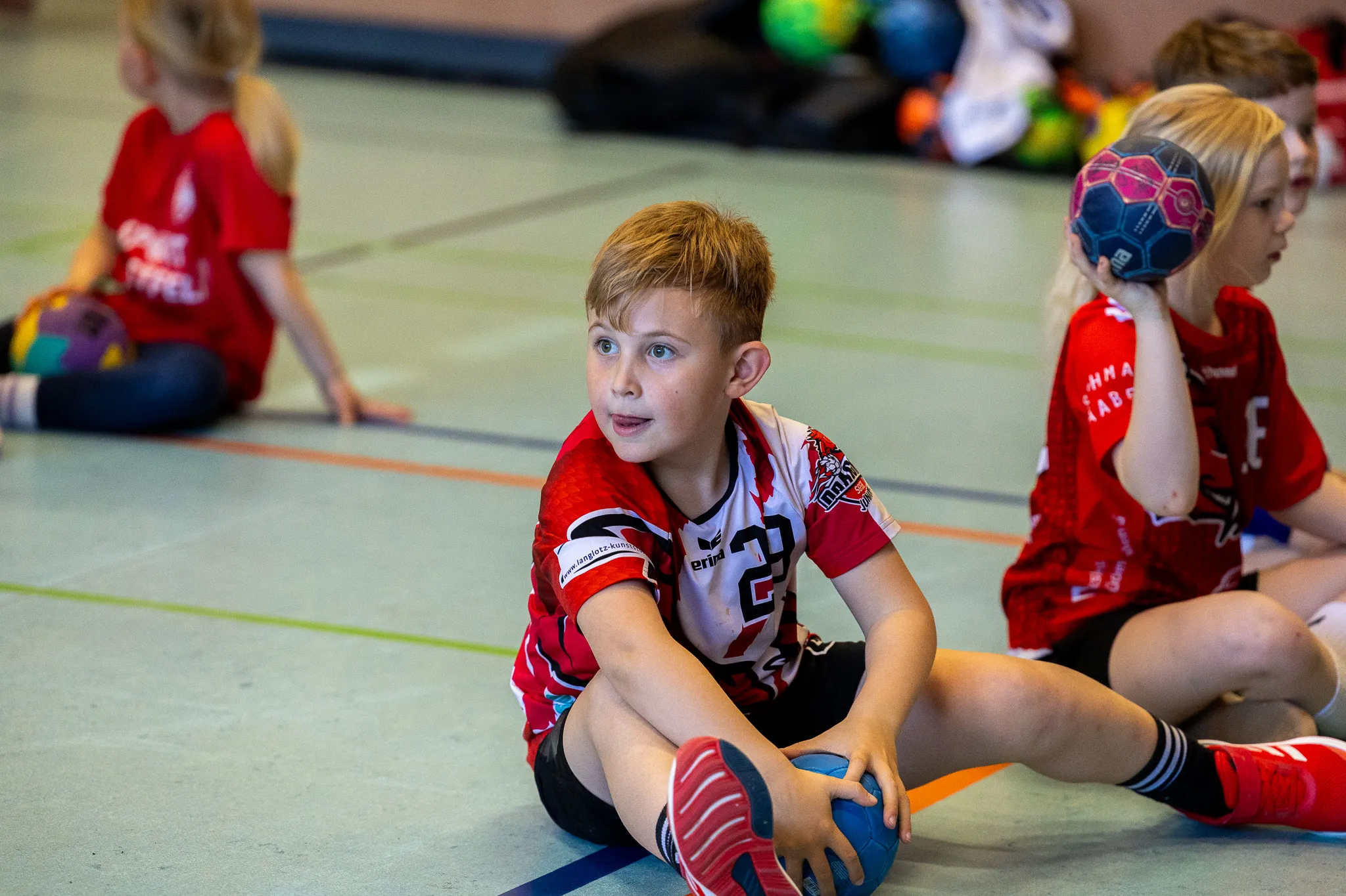 Junge sitzt beim Handballspiel auf dem Boden und hält einen Ball, während andere Kinder im Hintergrund spielen.