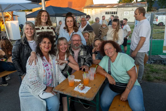 Eine Gruppe lächelnder Gäste posiert im Freien um einen Holztisch mit Getränken bei der Q-Stall-Party.