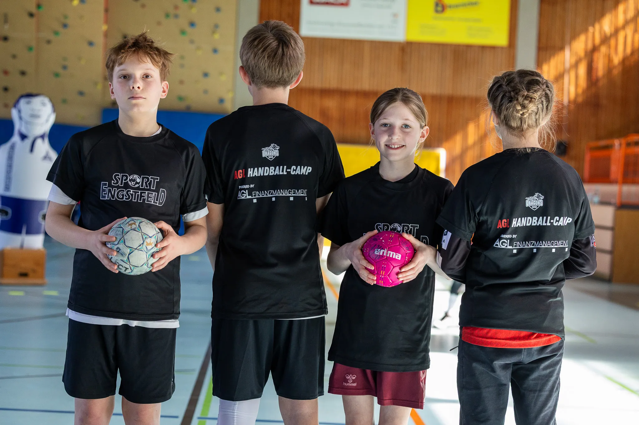 Zwei jugendliche Handballspieler mit Bällen und Trainingsanzügen mit Aufdrucken von Sponsoren stehen in einer Sporthalle.