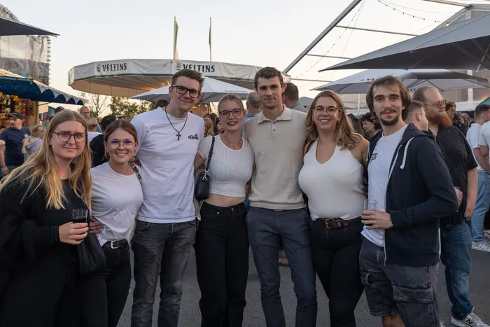 Eine Gruppe junger Menschen posiert lächelnd bei der Q-Stall-Party im Freien vor Sonnenschirmen.