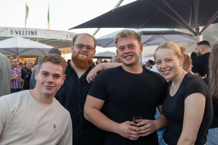 Vier lächelnde junge Menschen posieren bei der zehnten Q-Stall-Party unter Sonnenschirmen.