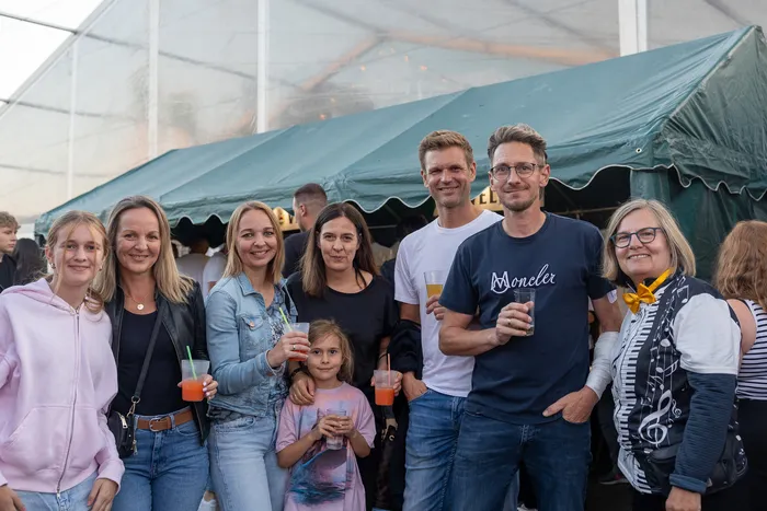 Eine Gruppe lächelnder Gäste mit Getränken posiert vor einem grünen Festzelt.