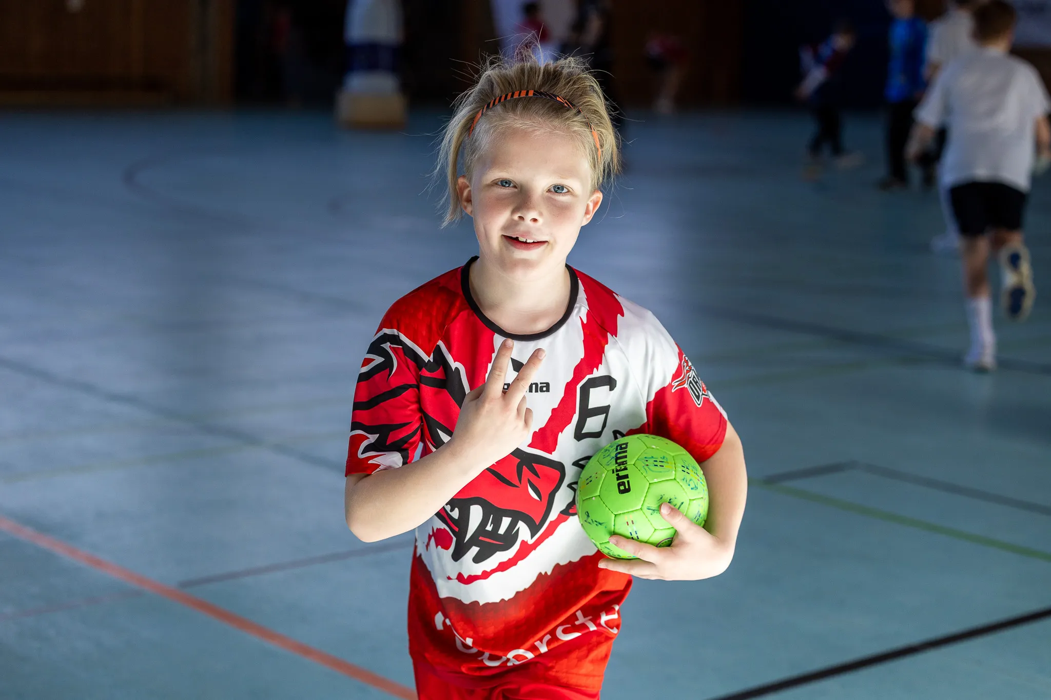 Ein junges Mädchen mit blonden Zöpfen hält einen grünen Handball in der Hand und lächelt in die Kamera. Sie trägt ein rotes Handballtrikot mit der Nummer 6.
