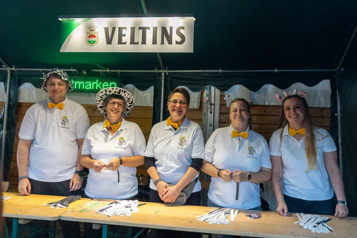 Fünf Personen in weißen Poloshirts und gelben Fliegen stehen hinter einem Tisch unter einem Veltins-Schild.