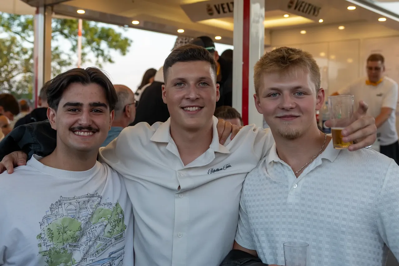 Drei junge Männer lächeln bei der Q-Stall-Party in Schalksmühle, einer hält ein Glas Bier.
