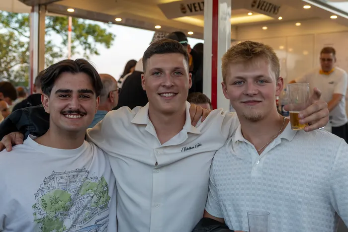 Drei junge Männer lächeln bei der Q-Stall-Party in Schalksmühle, einer hält ein Glas Bier.