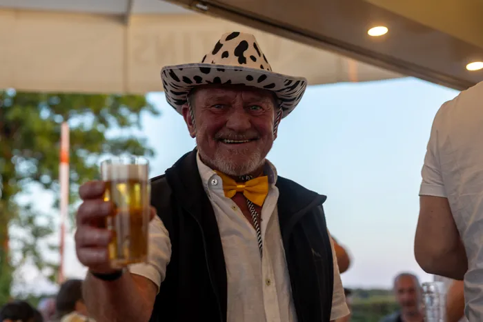 Ein lächelnder Mann mit gepunktetem Hut und gelber Fliege hebt ein Glas Bier zum Anstoßen.