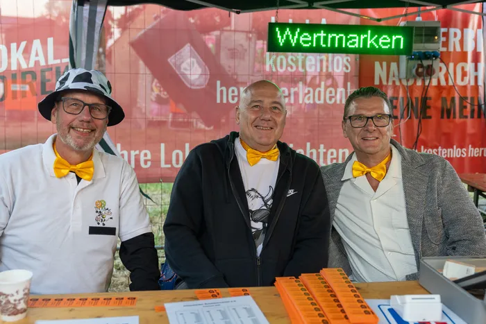 Drei lächelnde Männer mit gelben Fliegen stehen hinter einem Tisch vor einem grünen „Wertmarken“-Schild bei der Q-Stall-Party.