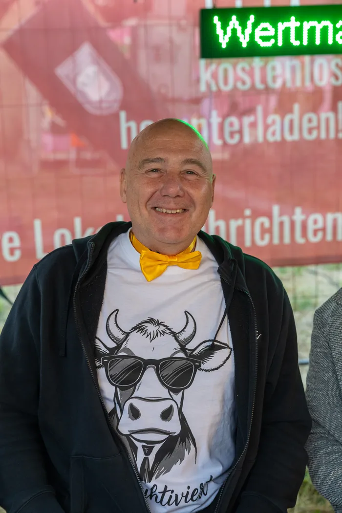 Ein lächelnder Mann mit gelber Fliege und einem T-Shirt mit Kuh-Motiv vor einem roten Banner.