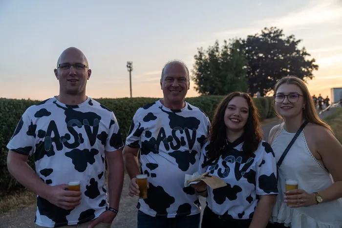 Vier lächelnde Personen bei der Q-Stall-Party, drei davon in weißen T-Shirts mit schwarzen Kuhflecken und dem Aufdruck „ASV“.