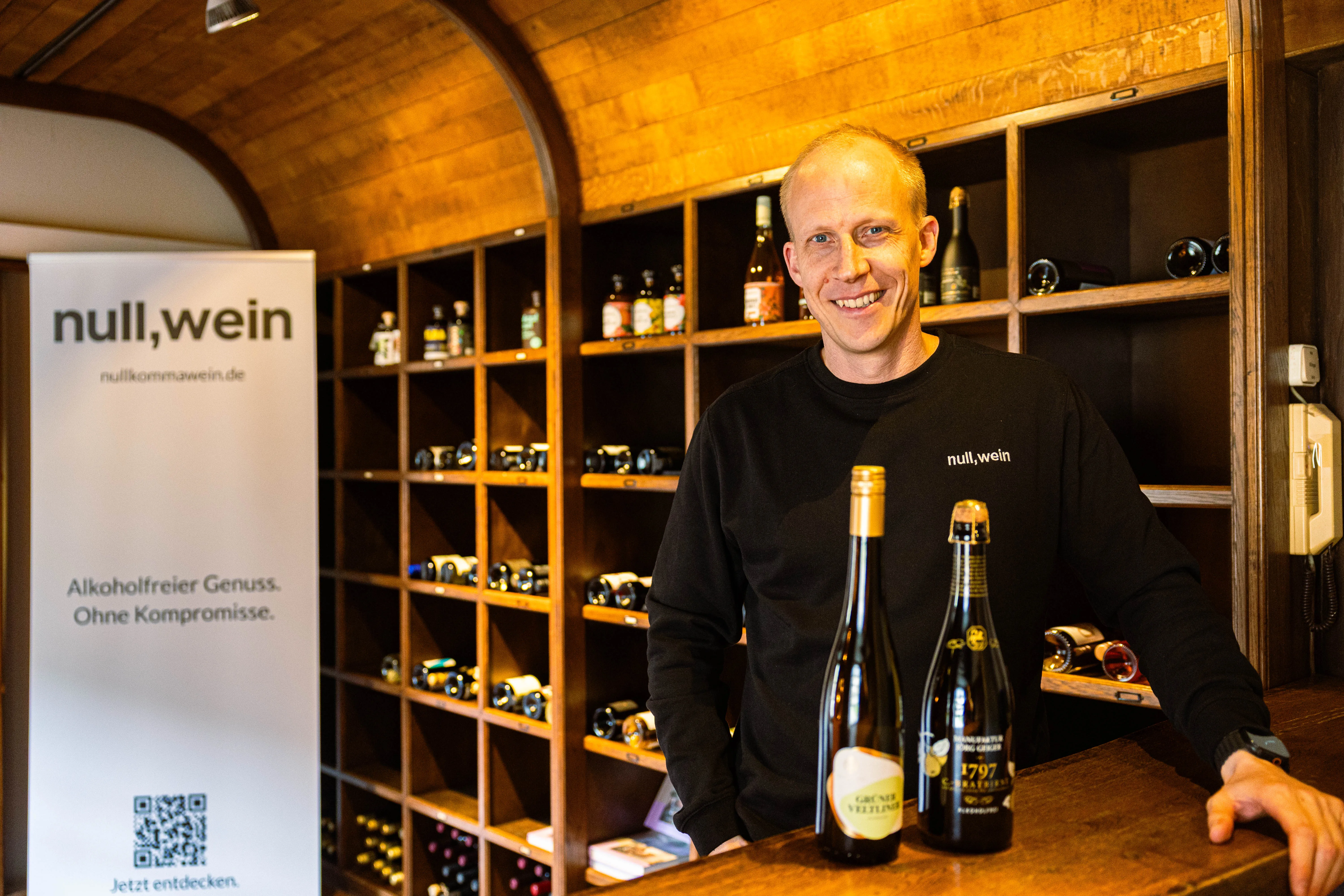 Hendrik Schellscheidt, Gründer von NullkommaWein, steht lächelnd vor Weinflaschen in einem Raum mit Weinfächern im Hintergrund. Links ein Banner mit dem NullkommaWein-Logo und dem Slogan „Alkoholfreier Genuss. Ohne Kompromisse.“
