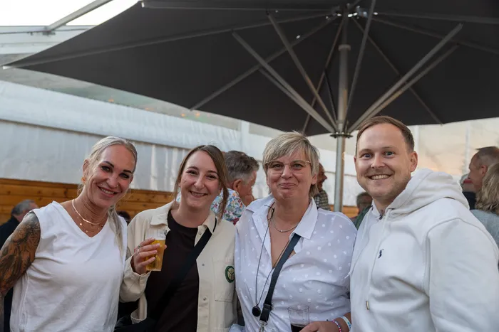 Vier lächelnde Gäste stehen unter einem großen grauen Sonnenschirm bei der Q-Stall-Party.