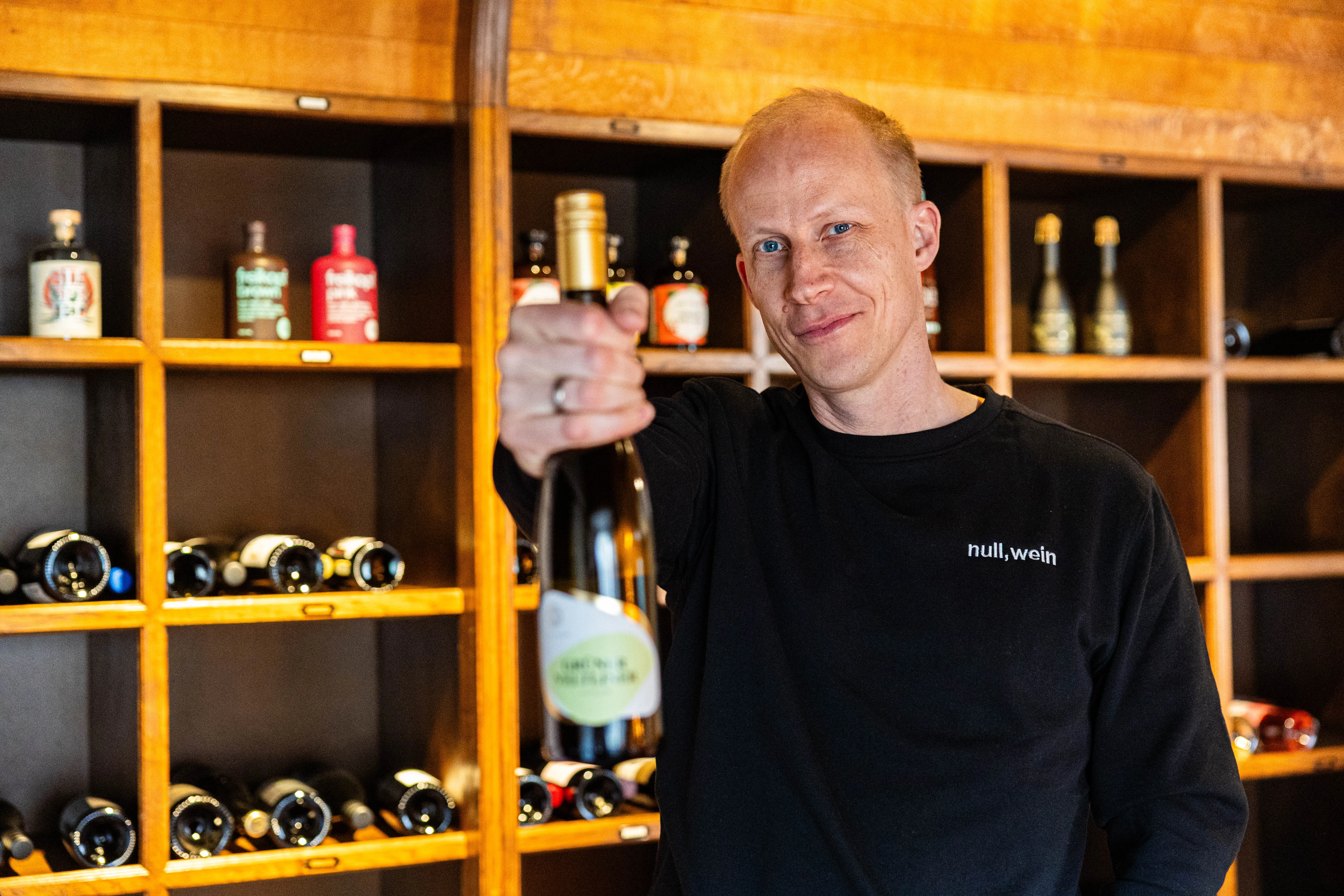 Hendrik Schellscheidt, Gründer von NullkommaWein, hält eine Weinflasche vor einem Regal mit weiteren Weinflaschen im Hintergrund. Er steht in einem Raum im Schloss Neuenhof, der für Verkostungen genutzt wird.