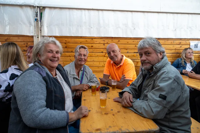 Eine Gruppe lächelnder Gäste sitzt an einem Holztisch mit Biergläsern bei der Q-Stall-Party.