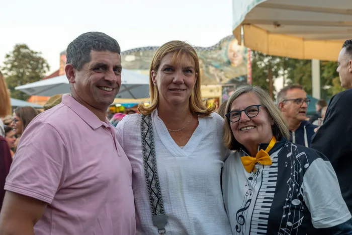 Drei lächelnde Gäste, darunter ein Mann in einem rosa Poloshirt und eine Frau mit gelber Fliege, posieren bei der Q-Stall-Party.