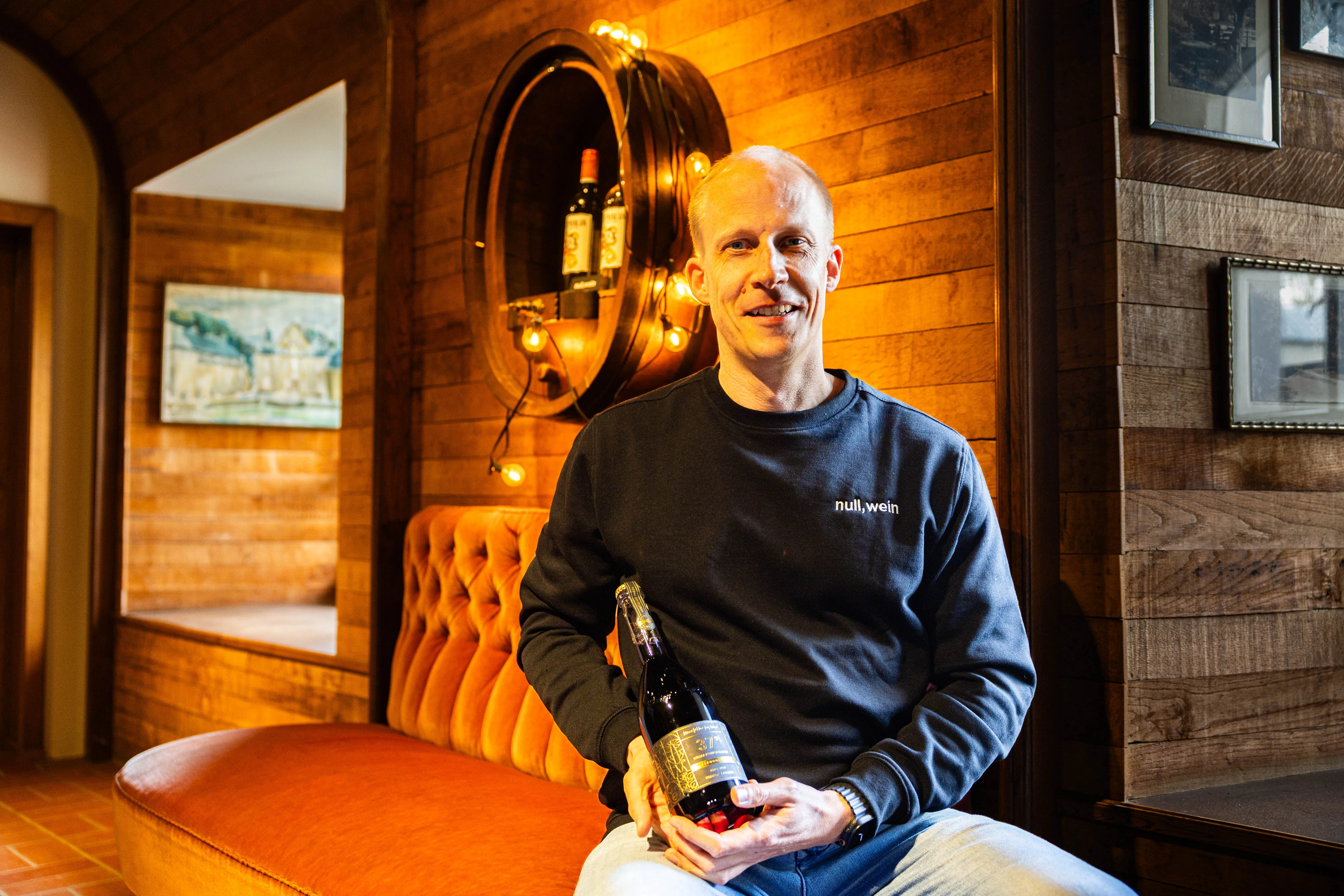 Hendrik Schellscheidt, Gründer von NullkommaWein, sitzt lächelnd mit zwei Weinflaschen in einem Raum mit orangefarbenem Sofa und Holzvertäfelung.