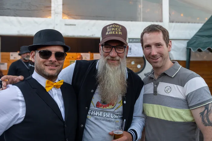 Drei lächelnde Männer, einer mit Bowler-Hut und gelber Fliege sowie einer mit langem Bart und einem Bierglas, posieren bei der zehnten Q-Stall-Party.