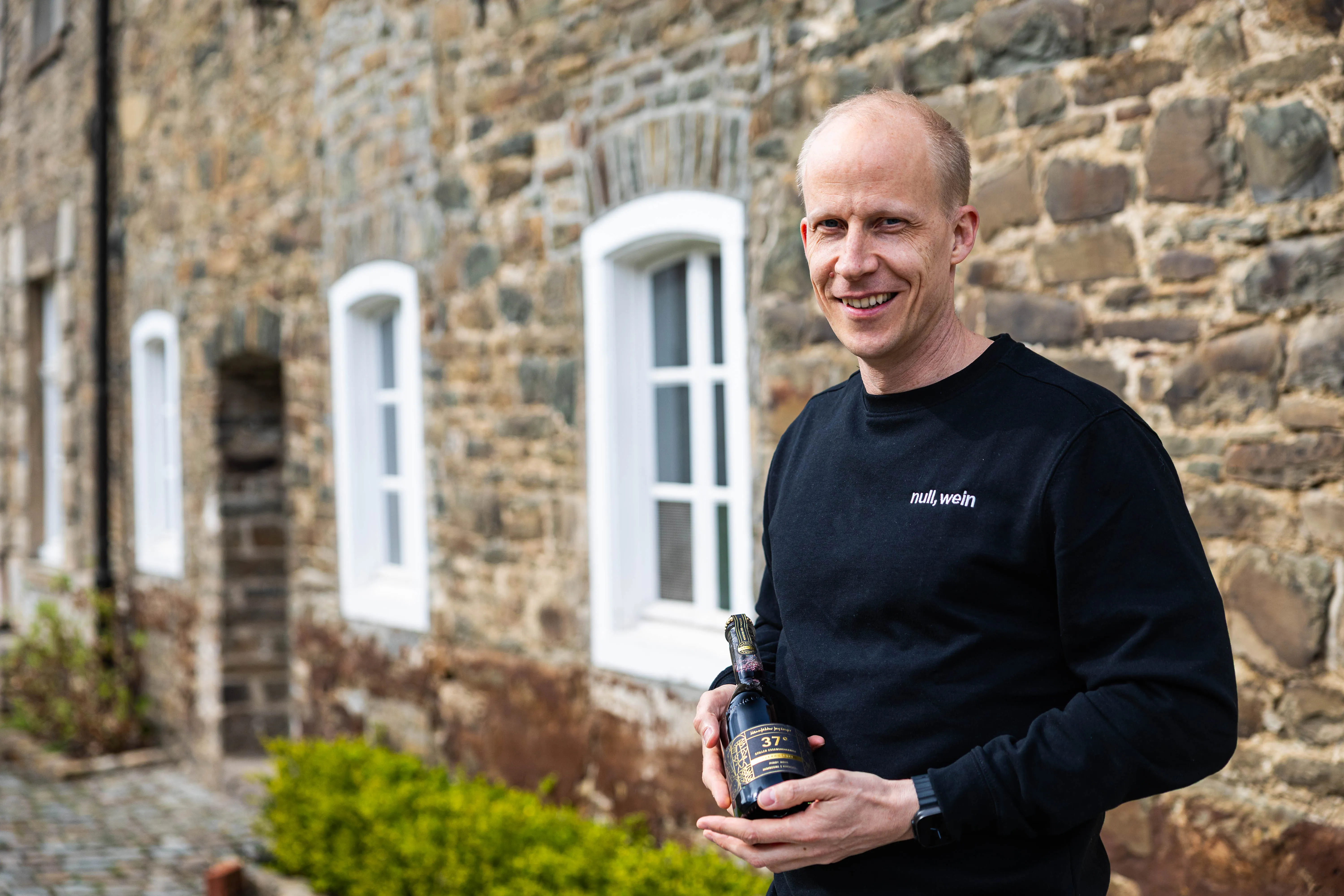 Hendrik Schellscheidt, Gründer von NullkommaWein, lächelt mit einer Flasche Wein vor dem historischen Schloss Neuenhof in Lüdenscheid.