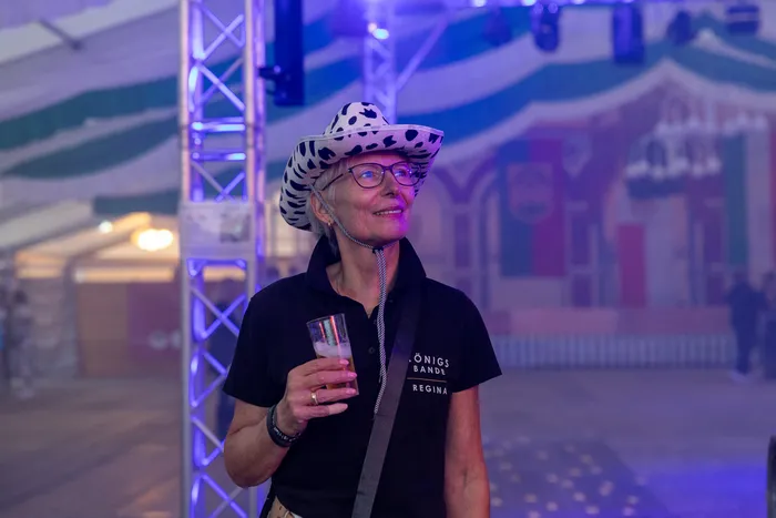 Eine lächelnde Frau mit Kuhprint-Hut und Brille hält ein Glas Bier bei einer Party in einem blau beleuchteten Zelt.