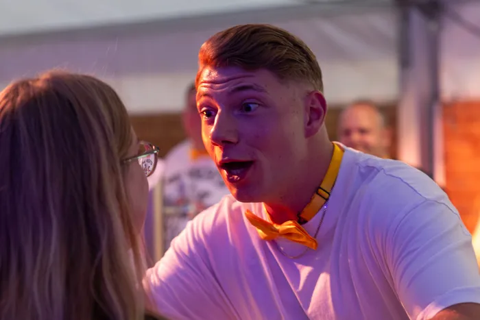 Ein junger Mann mit gelber Fliege und weit geöffnetem Mund blickt eine Frau auf einer Party an.