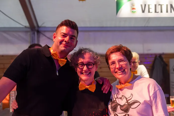 Drei lächelnde Gäste mit orangefarbenen Fliegen bei der zehnten Q-Stall-Party in Schalksmühle.