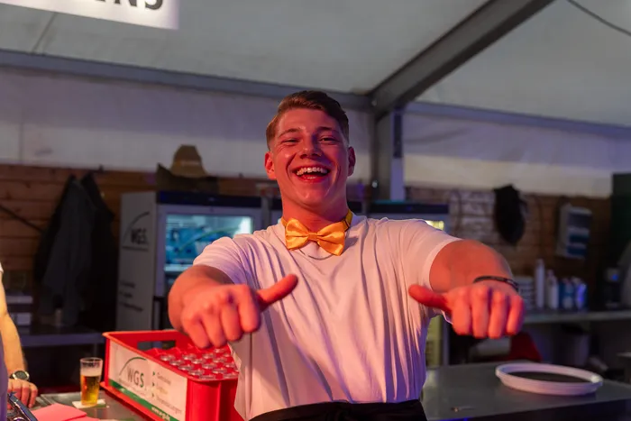Ein lachender junger Mann in einem weißen T-Shirt und einer gelben Fliege zeigt mit beiden Daumen nach vorne.