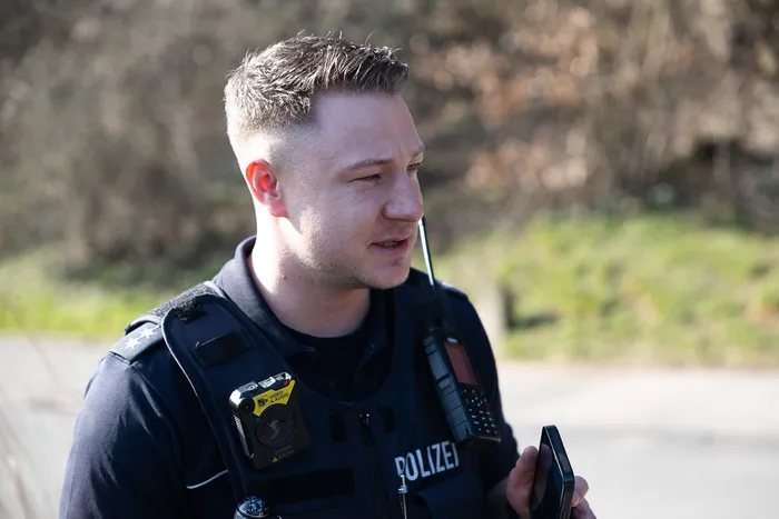Ein Polizist in Uniform blickt zur Seite, während er ein Funkgerät hält. Am Revers ist eine Bodycam befestigt.